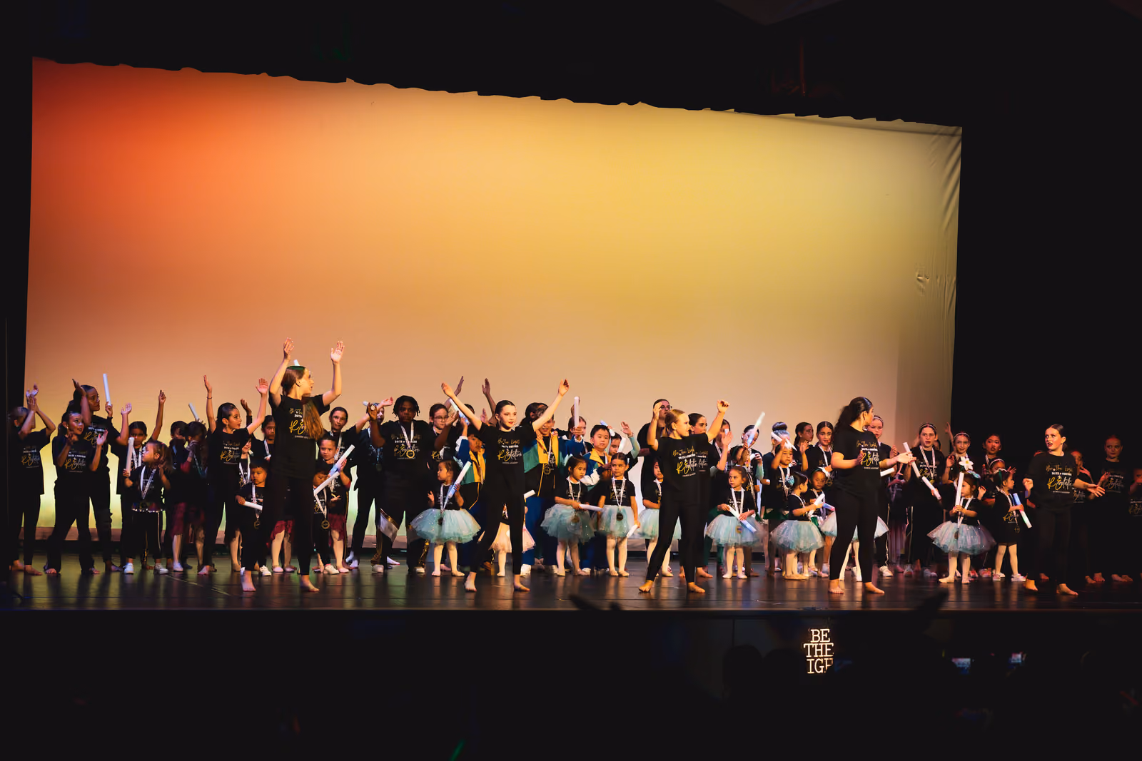 Children and teens on stage during a recital finale, some in tutus and others in black shirts with gold writing.