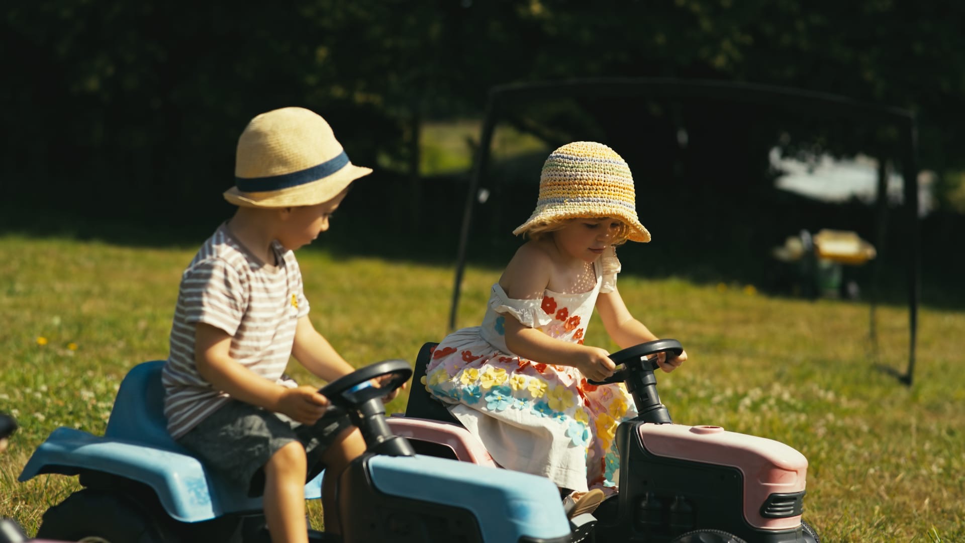 Two small children in sun hats riding toy tractors across the grass