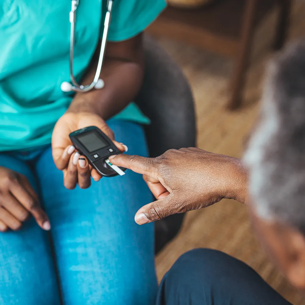A nurse in scrubs performing a fingerstick blood test for a seated patient using a handheld meter