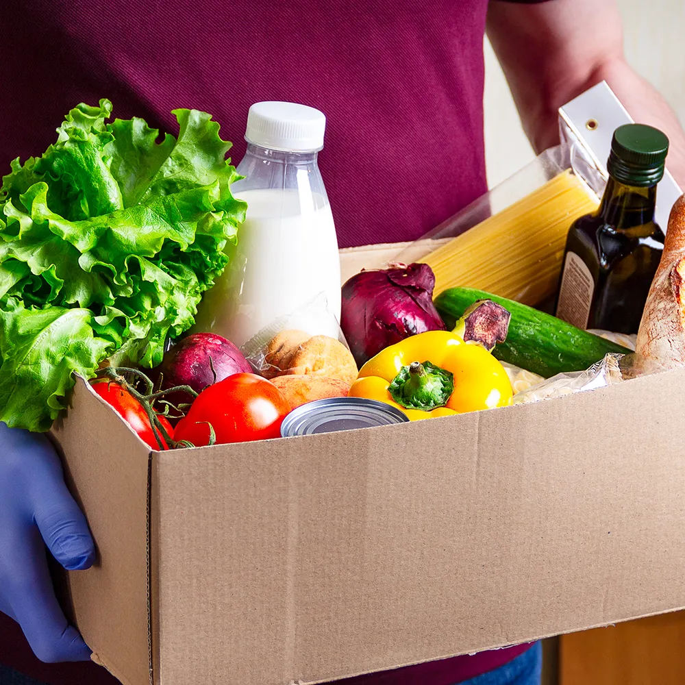 A volunteer carrying a box of groceries including fresh vegetables, bread and milk