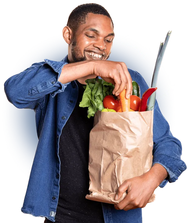 A smiling man carrying a paper grocery bag filled with fresh lettuce, tomatoes, carrots and peppers