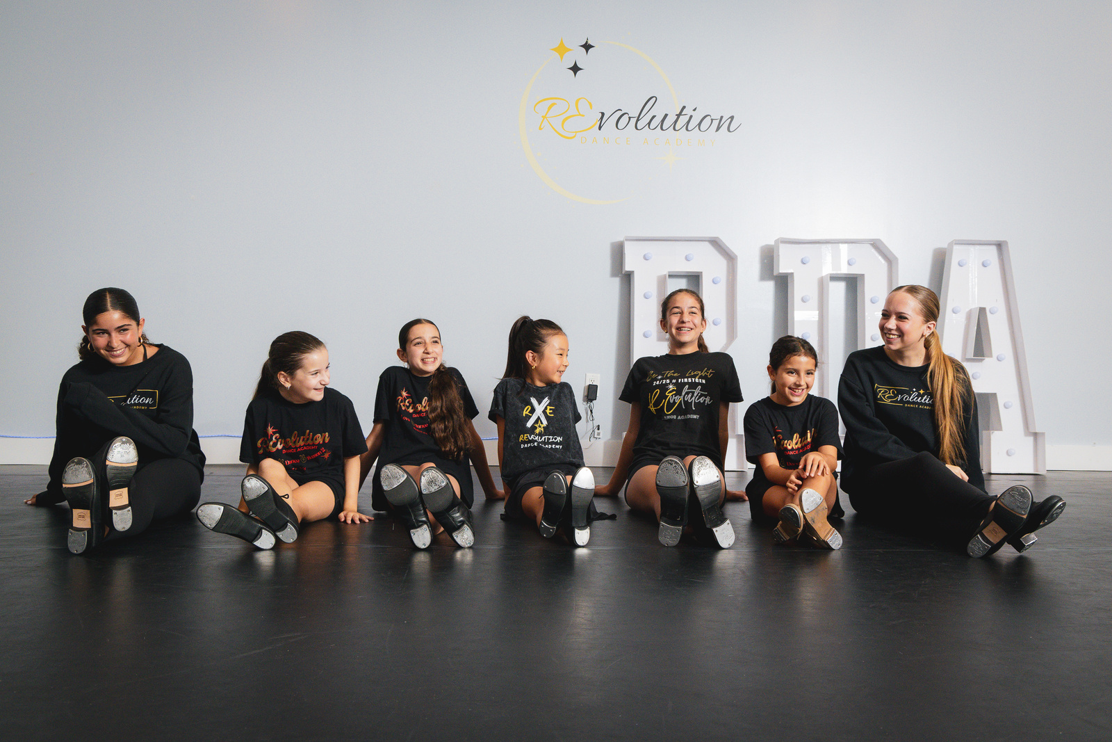 Tap students seated in a line showing off their tap shoes at Revolution Dance Academy