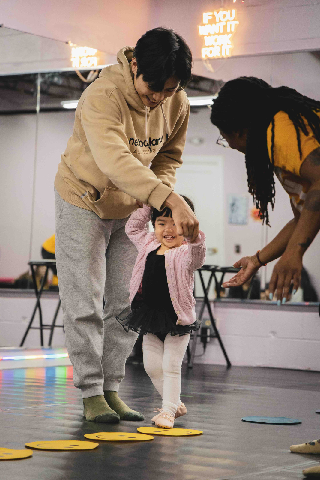 Adult helping a smiling toddler walk on yellow smiley face cutouts on the floor in a dance studio.