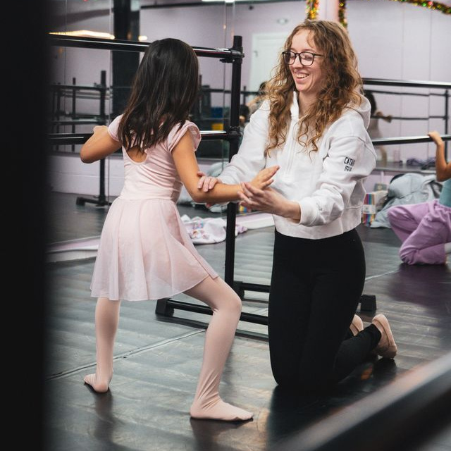 An RDA instructor kneeling to guide a young ballet student's arms during a private lesson in the Tenafly studio