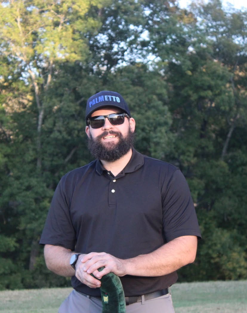 Man with a beard wearing a black polo shirt, sunglasses, and a blue cap with 'PALMETTO' written on it, standing outdoors in front of trees during daylight, holding a green object with both hands.