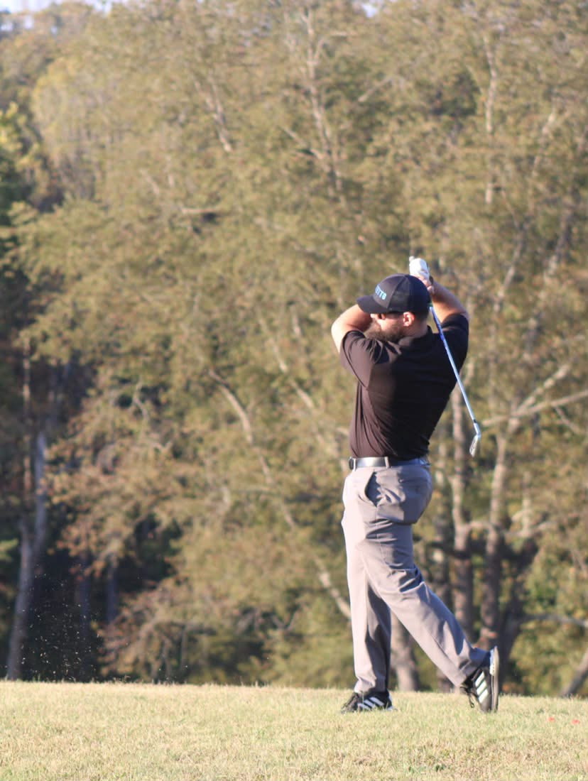 A man wearing a black shirt, gray pants, a black cap, and black golf shoes with white stripes is swinging a golf club on a grassy course with trees in the background.
