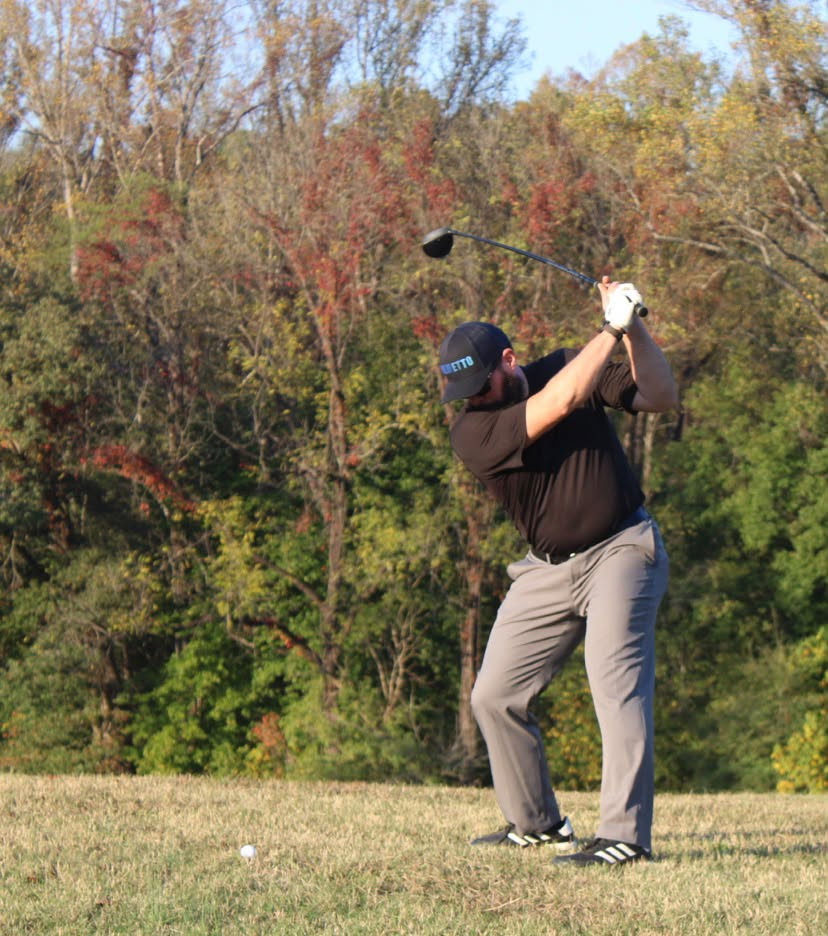 Golfer in a black shirt, gray pants, and a black cap is preparing to swing a golf club on a grassy area with trees showing autumn colors in the background.