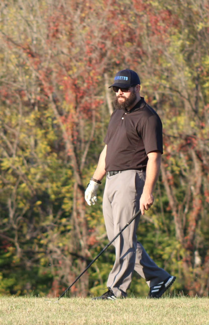 Man with sunglasses and a beard wearing a black polo, gray pants, black Adidas shoes, and a black cap walking on grass while holding a golf club, with autumn trees in the background.