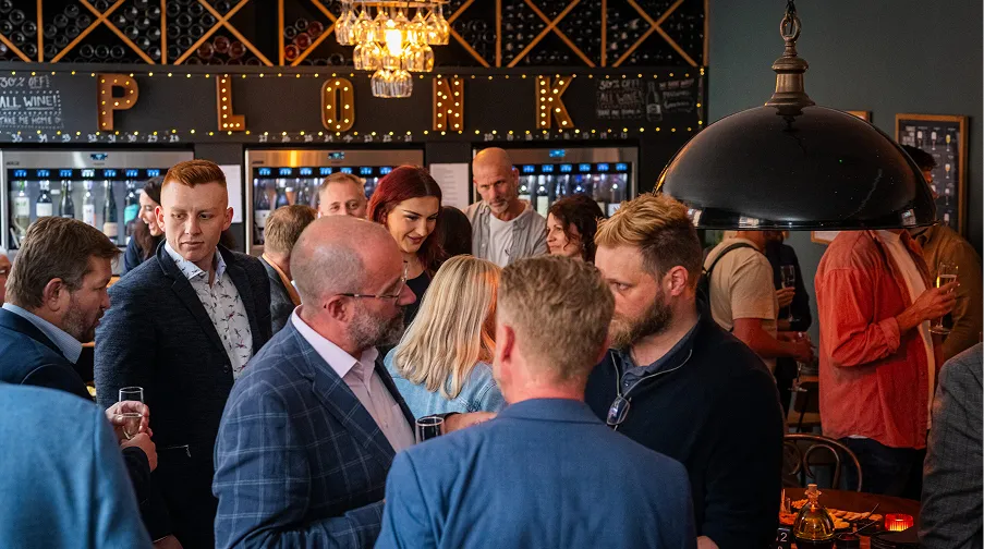 Group of people socializing in a bar with wine bottles and lit letters spelling PLONK in the background.