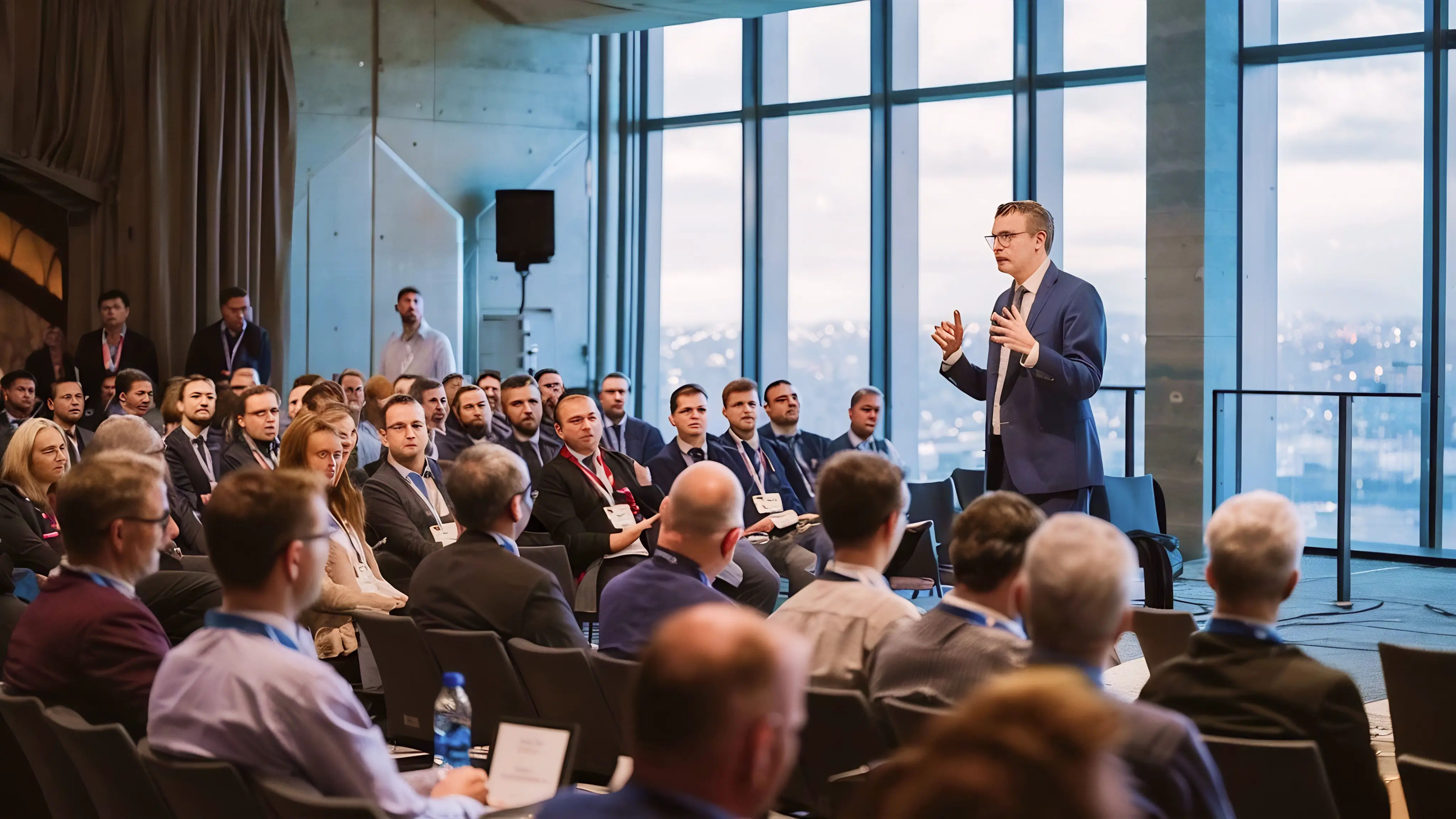Man in blue suit speaking to an attentive audience in a large room with tall windows.