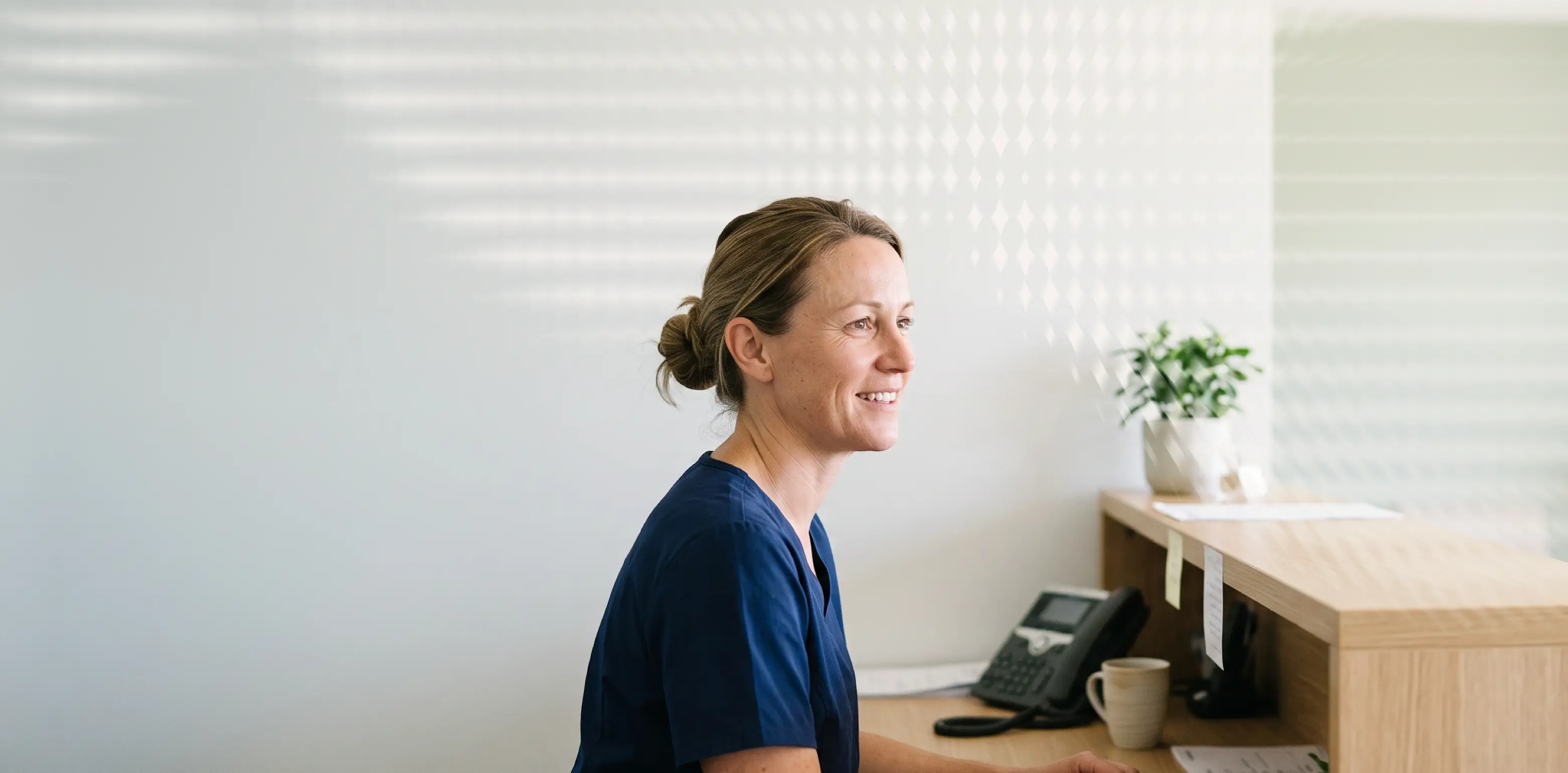 Woman in blue scrubs smiling at a desk with a phone and plant
