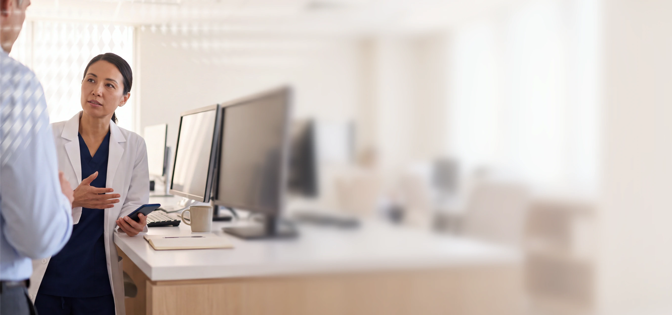 Medical professional in scrubs and lab coat discussing with colleague at computer desk in office setting