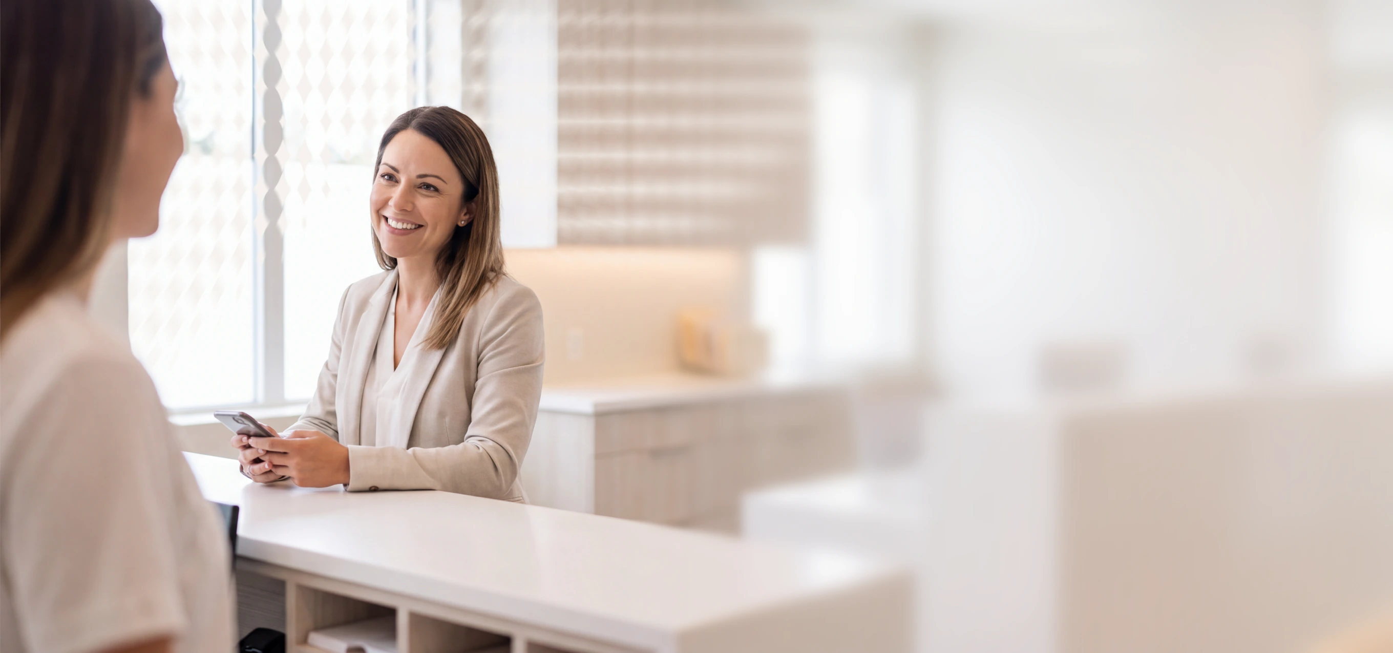 Woman in business attire smiling at a reception desk, holding a phone