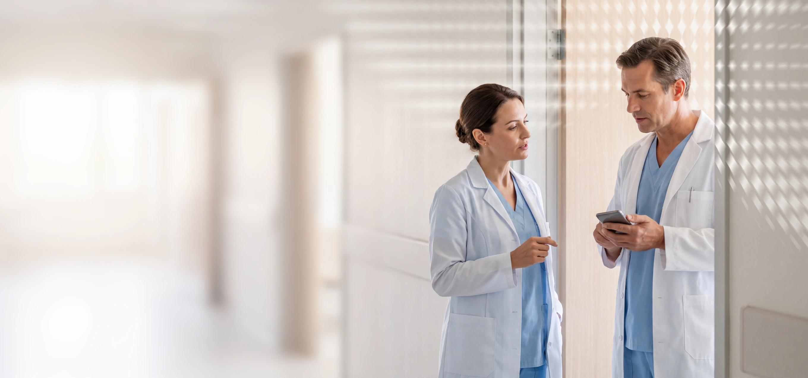Two healthcare professionals in scrubs and lab coats standing in a hallway, one holding a tablet