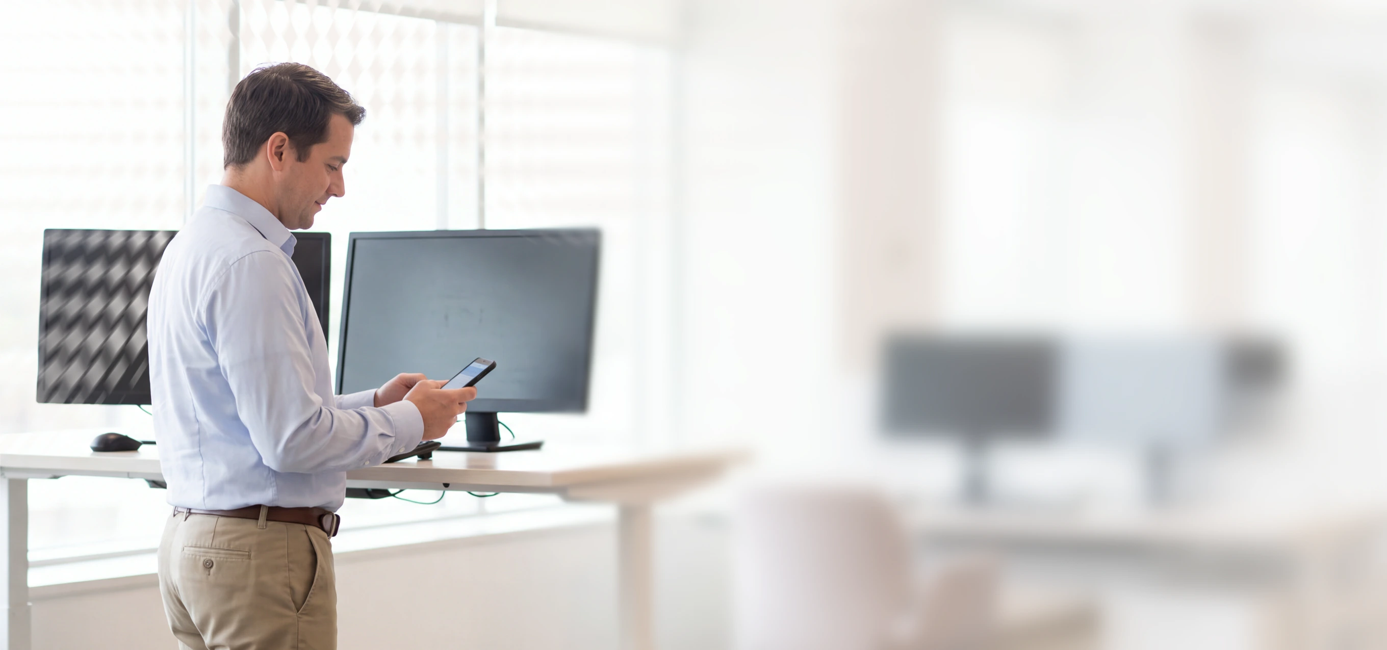 Man in business attire using a smartphone at a standing desk with multiple monitors