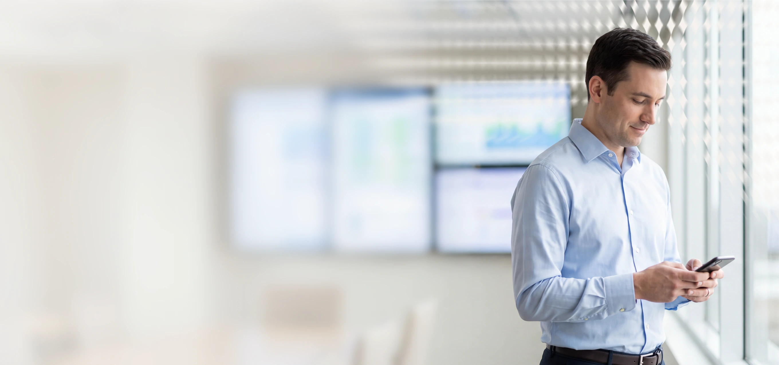Man in light blue shirt using a smartphone in an office setting with large windows and a digital display in the background