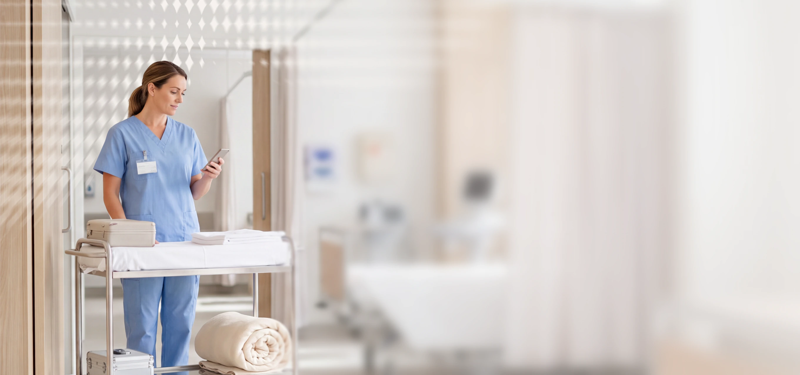 Nurse in scrubs using a phone in a hospital hallway with medical supplies on a trolley