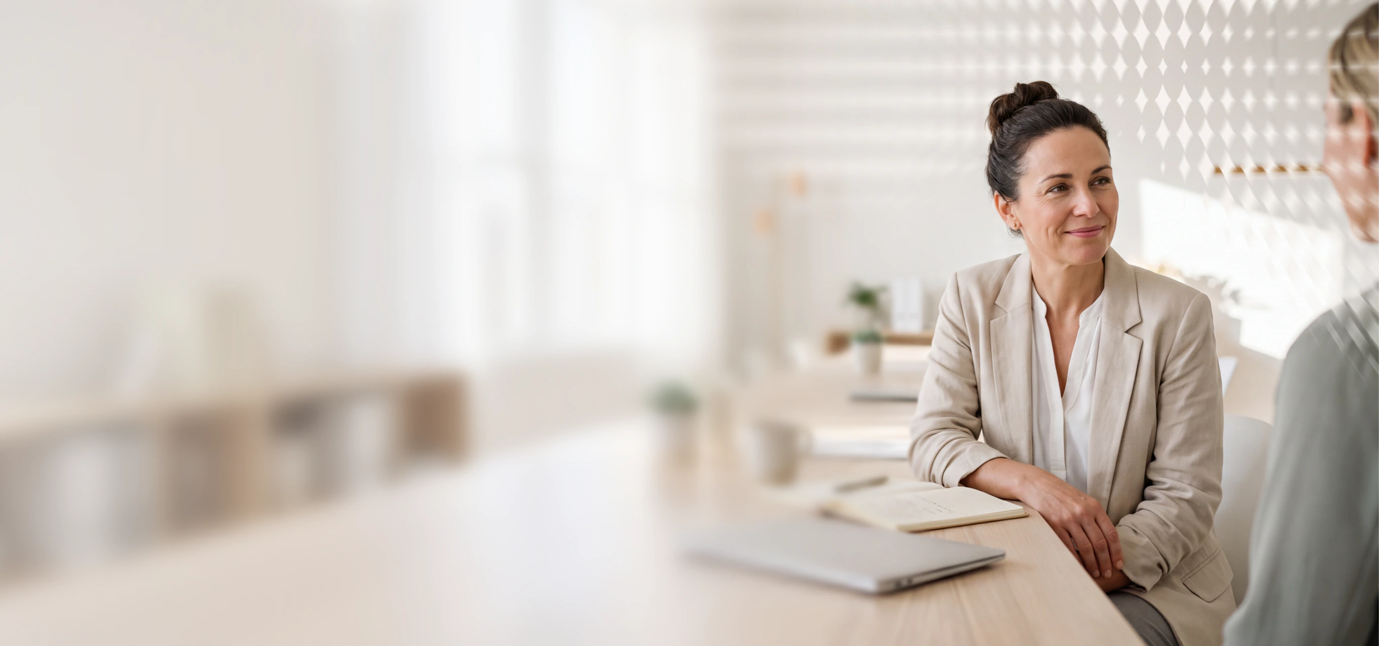 Professional woman in business attire seated at a desk with a laptop, engaging in a conversation