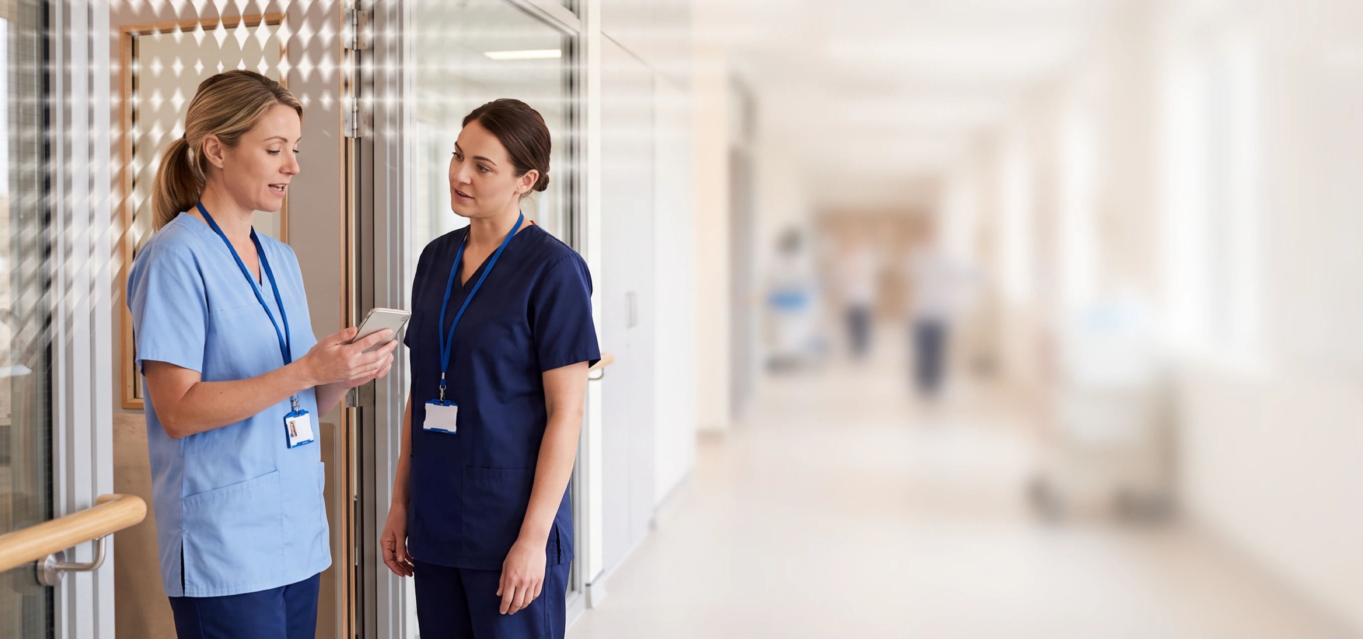 Two healthcare professionals in scrubs standing in a hallway, one holding a device