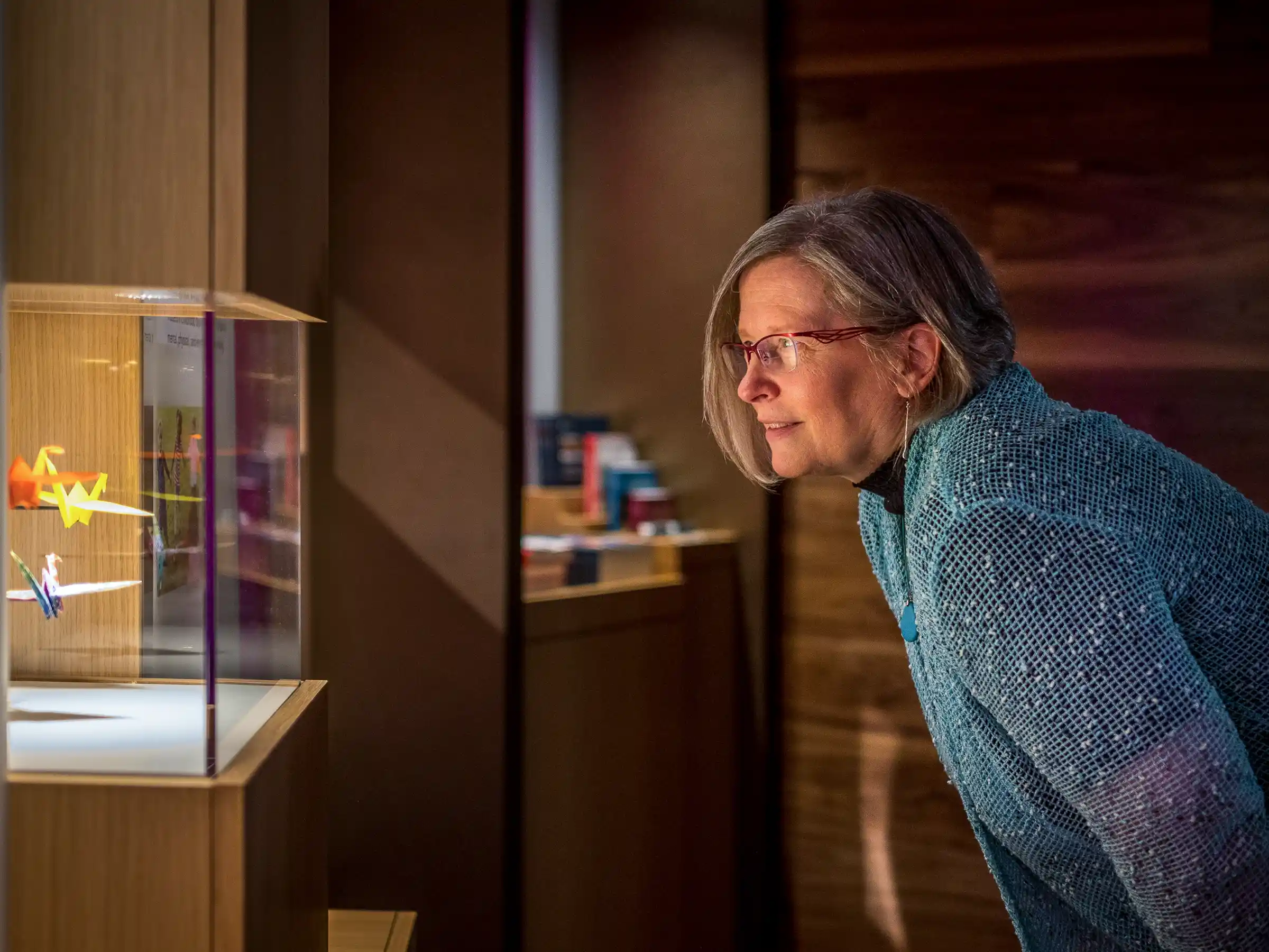 A woman wearing glasses and a blue textured sweater leaning forward to closely observe colorful origami cranes displayed inside a glass case in a warmly lit indoor setting.