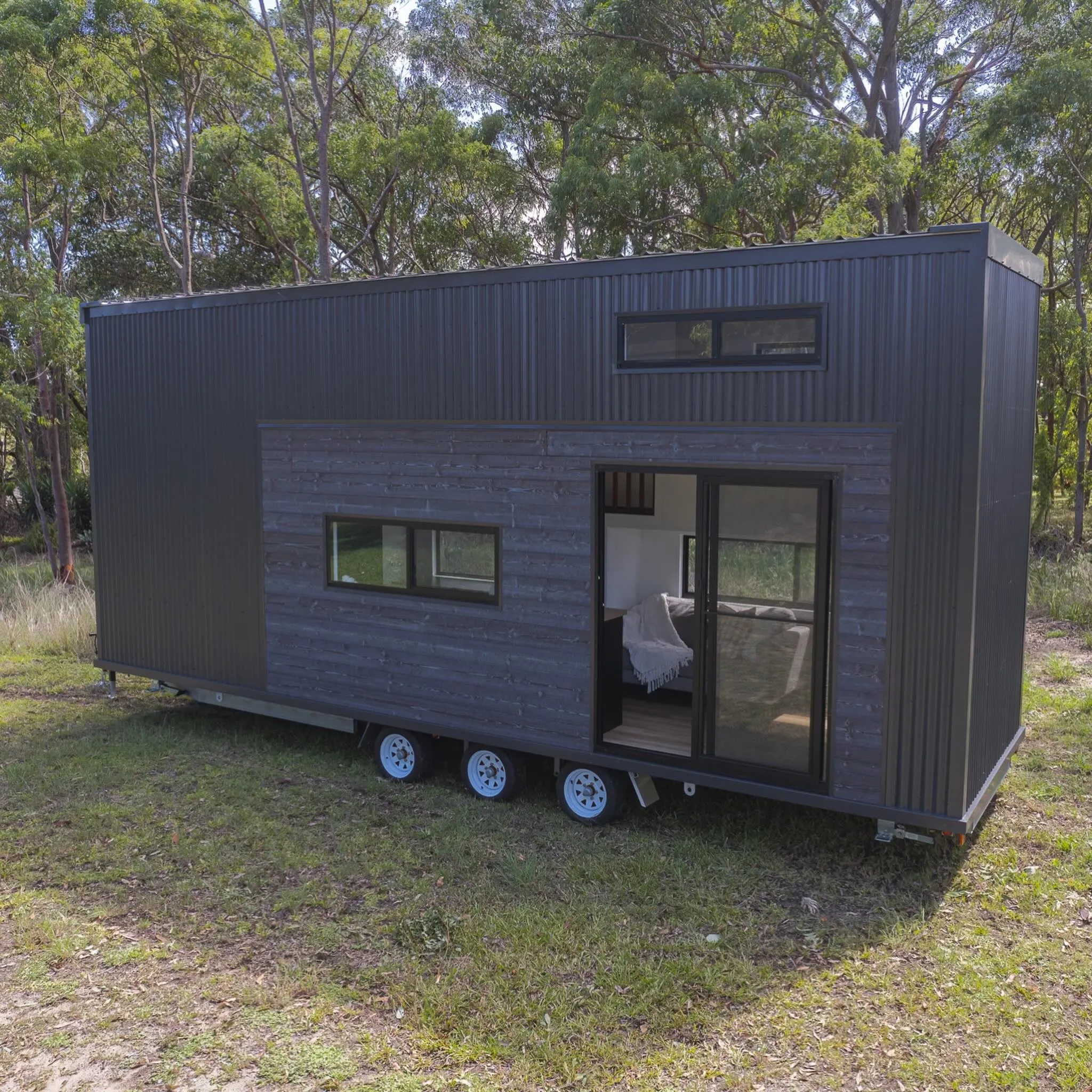 A modern tiny house on wheels with dark vertical and horizontal siding, large sliding glass door, and rectangular windows, set in a grassy area with trees around.