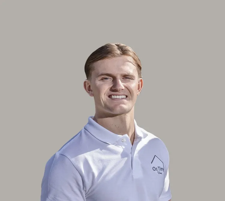 Young man with short light brown hair wearing a white polo shirt with a small logo on the chest, smiling against a plain gray background.