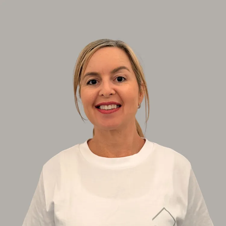 Smiling woman with blonde hair tied back wearing a white t-shirt against a plain gray background.