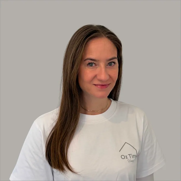 Woman with long brown hair wearing a white t-shirt with a small logo that says 'Oz Tiny Homes' on a plain light grey background.