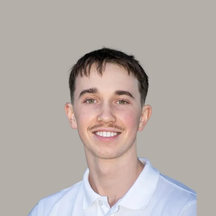 Portrait of a young man with light skin, short dark hair, a thin mustache, and green eyes, smiling and wearing a white collared shirt against a plain beige background.
