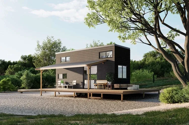 Modern tiny house with dark exterior walls on a raised wooden platform surrounded by green trees and bushes under a clear sky.