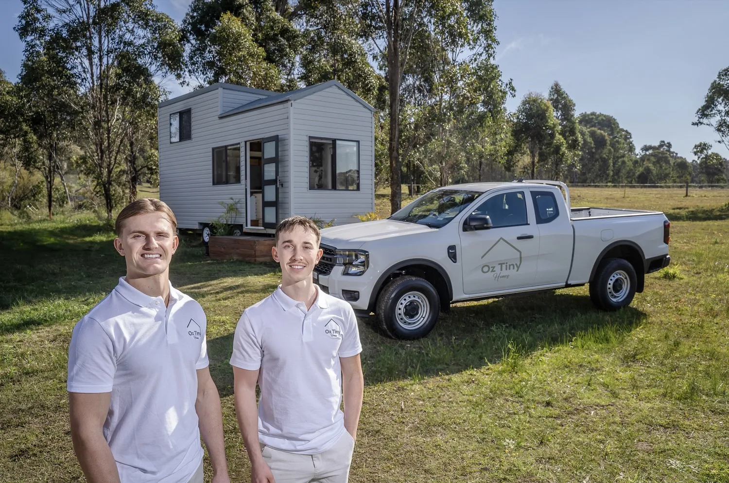 Two men wearing white polo shirts with Oz Tiny Homes logo standing in front of a tiny house and a white pickup truck with the same logo, all set on grass with trees in the background under a clear sky.