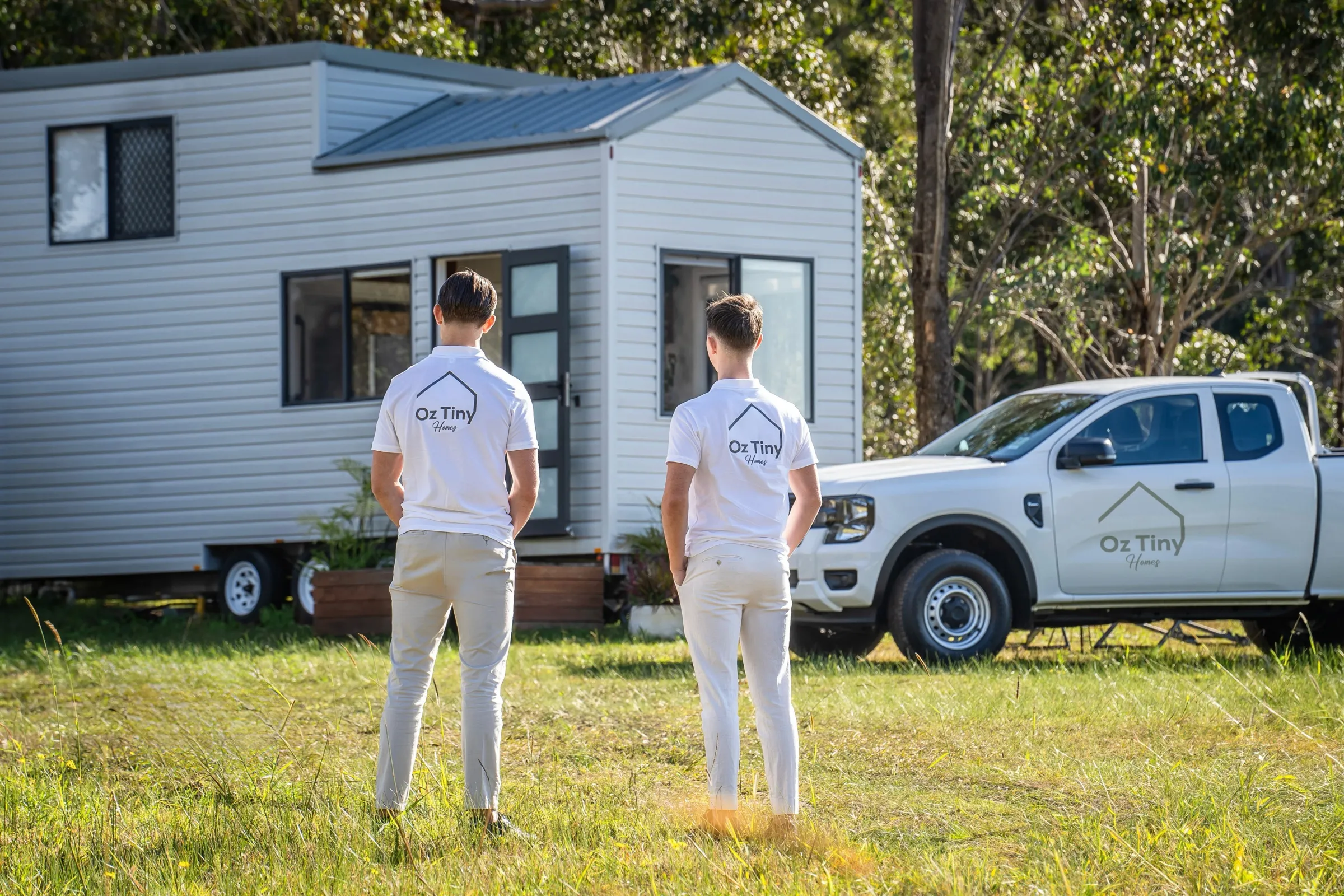 Two men wearing white shirts with Oz Tiny Homes logo stand on grass facing a small white tiny house and a white pickup truck with the same logo, surrounded by trees.