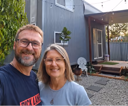 Smiling middle-aged man and woman wearing glasses take a selfie outdoors in front of a modern gray metal-sided house with a wooden deck, stepping stones in gravel, potted plants, and string lights overhead.