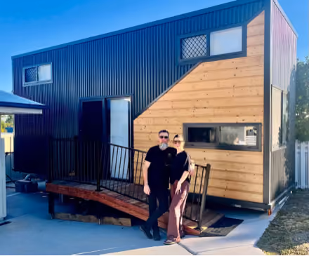 A couple wearing sunglasses stands in front of a modern tiny house with black corrugated metal and natural wood siding, a small ramp, and three horizontal windows.