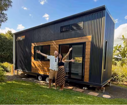 A smiling man and woman with arms outstretched stand in front of a modern tiny house on wheels with black siding and wooden accents, set on a green lawn with trees and blue sky in the background.