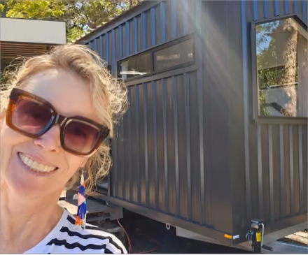 Smiling woman wearing large square sunglasses and colorful beaded earrings taking a selfie outdoors near a dark gray metal tiny house on wheels.