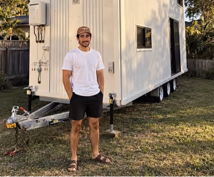 A man wearing a white t-shirt, black shorts, brown cap, and brown sandals stands on grass beside a white tiny house on wheels with three visible tires and several small windows.