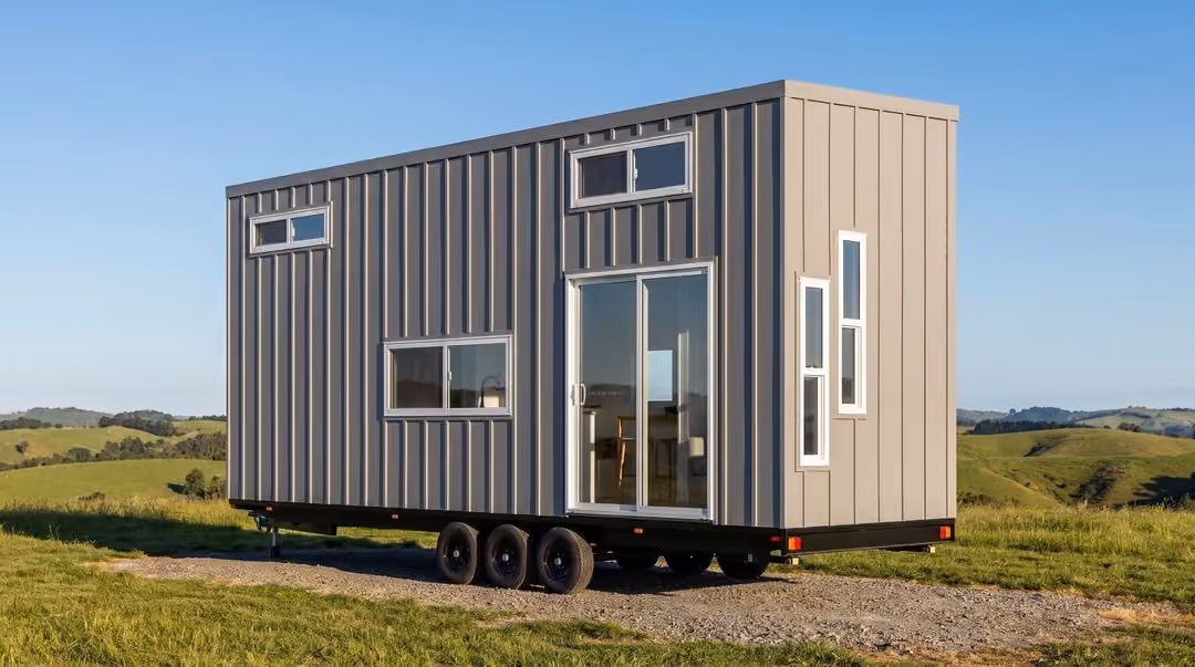 Modern tiny house on wheels with vertical panel siding, multiple rectangular windows, and a sliding glass door, set in a grassy open landscape under a clear blue sky.