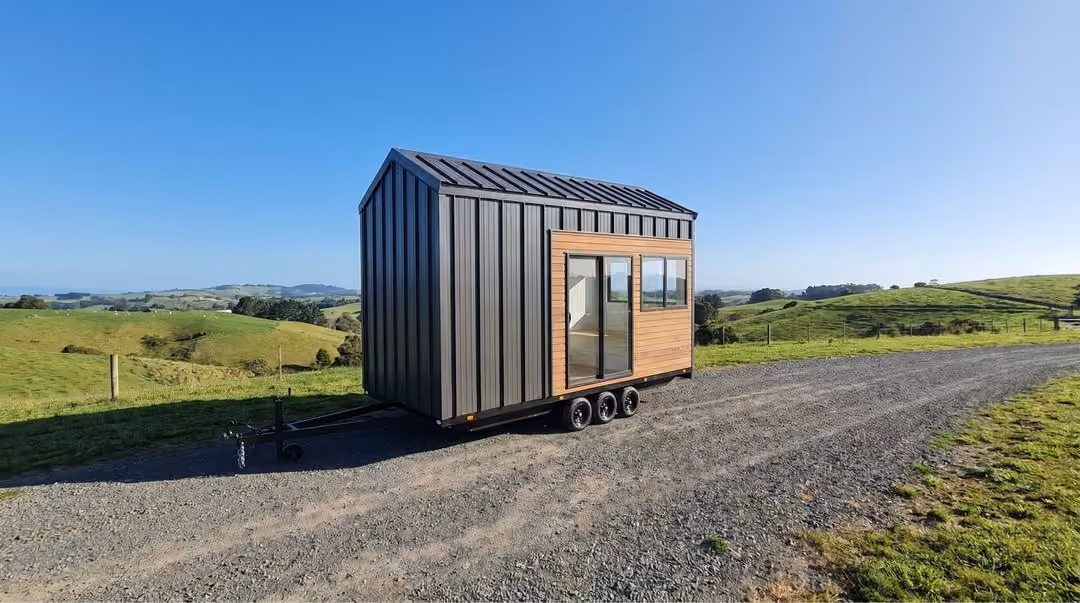 Modern tiny house on wheels parked on a gravel road surrounded by green rolling hills under a clear blue sky.