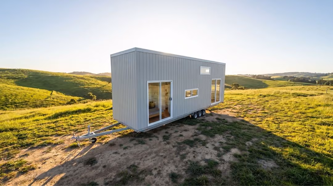 Modern tiny house on wheels with gray siding parked on a dirt patch in a green grassy field under a clear sky at sunset.