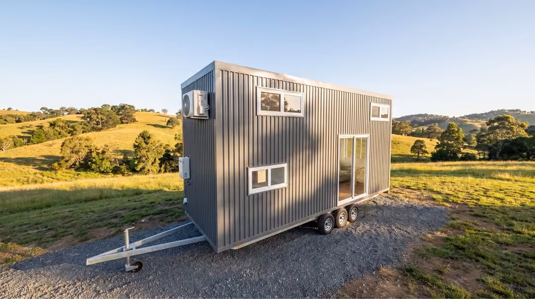 Modern tiny house on wheels with dark vertical siding, multiple rectangular windows, and an air conditioning unit, parked on gravel in a sunny rural area with grassy hills and scattered trees.