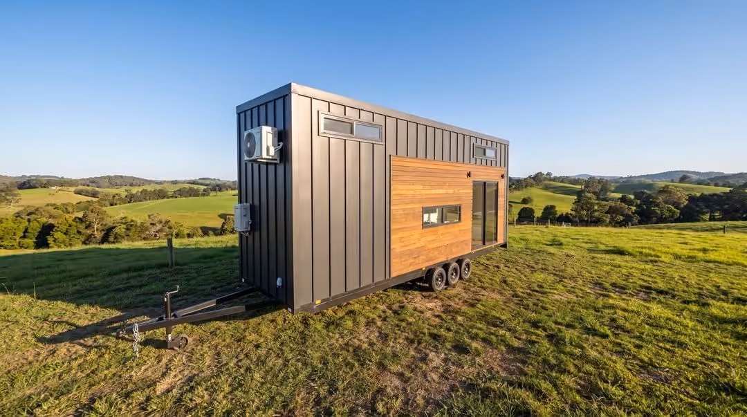 Modern tiny house on wheels with black metal and wooden panel exterior, parked on grassy countryside under clear blue sky.