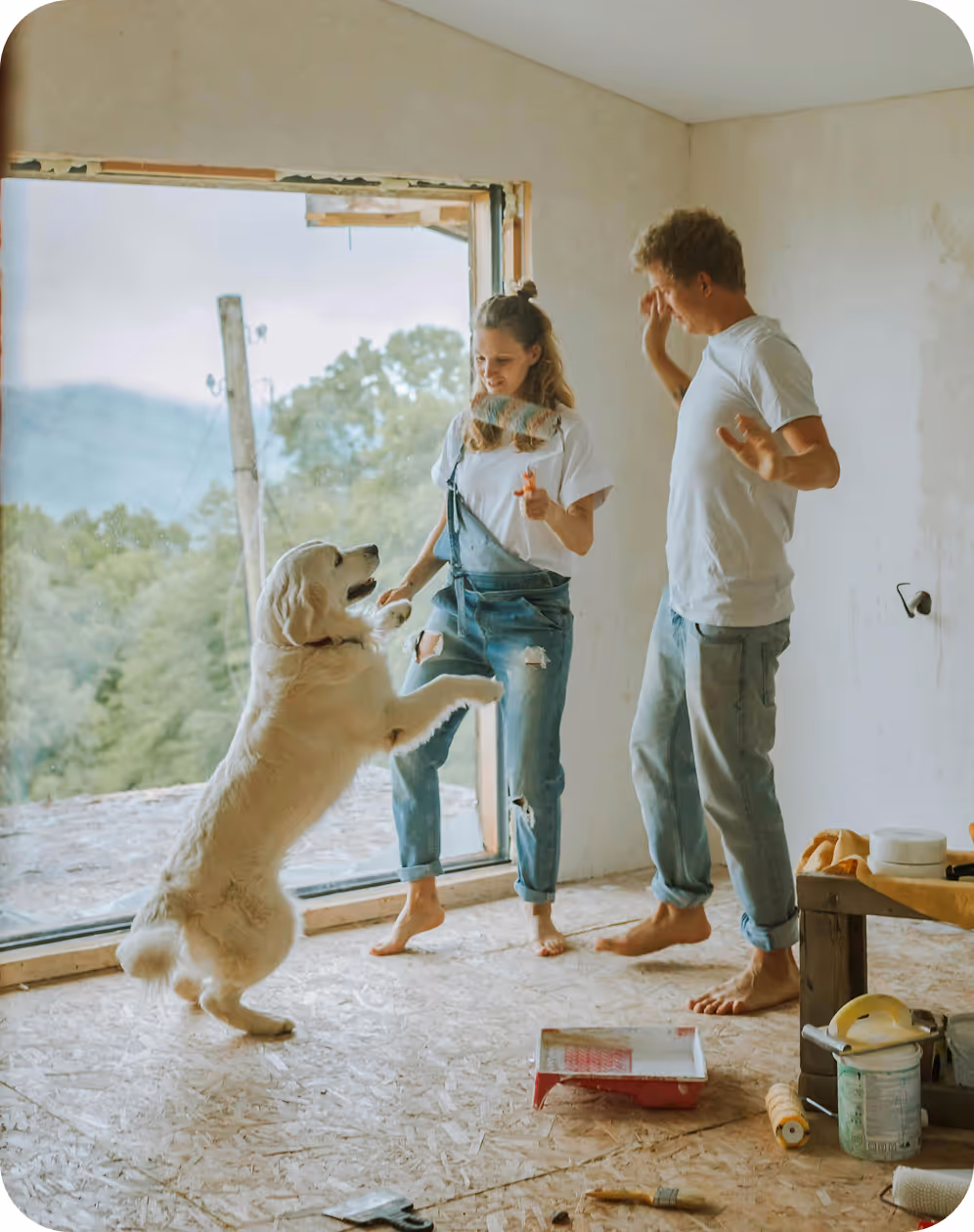 Young couple playing with a golden retriever inside a partially renovated room with painting supplies on the floor.