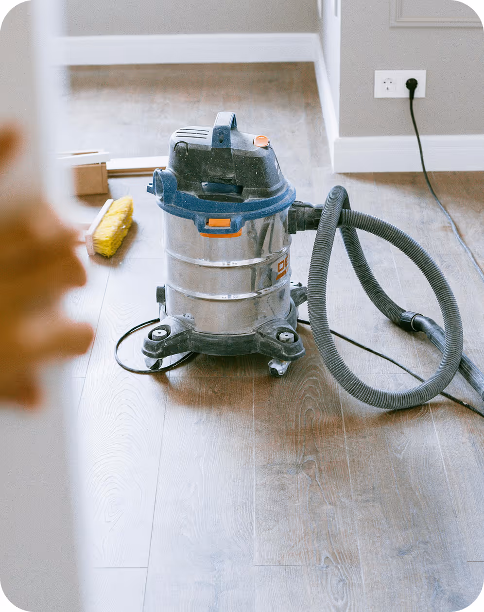 Industrial vacuum cleaner on wooden floor near a wall outlet with a yellow brush in the background.
