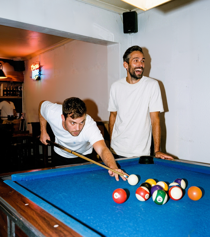 Two men playing pool; one aims the cue ball while the other watches smiling at the table.