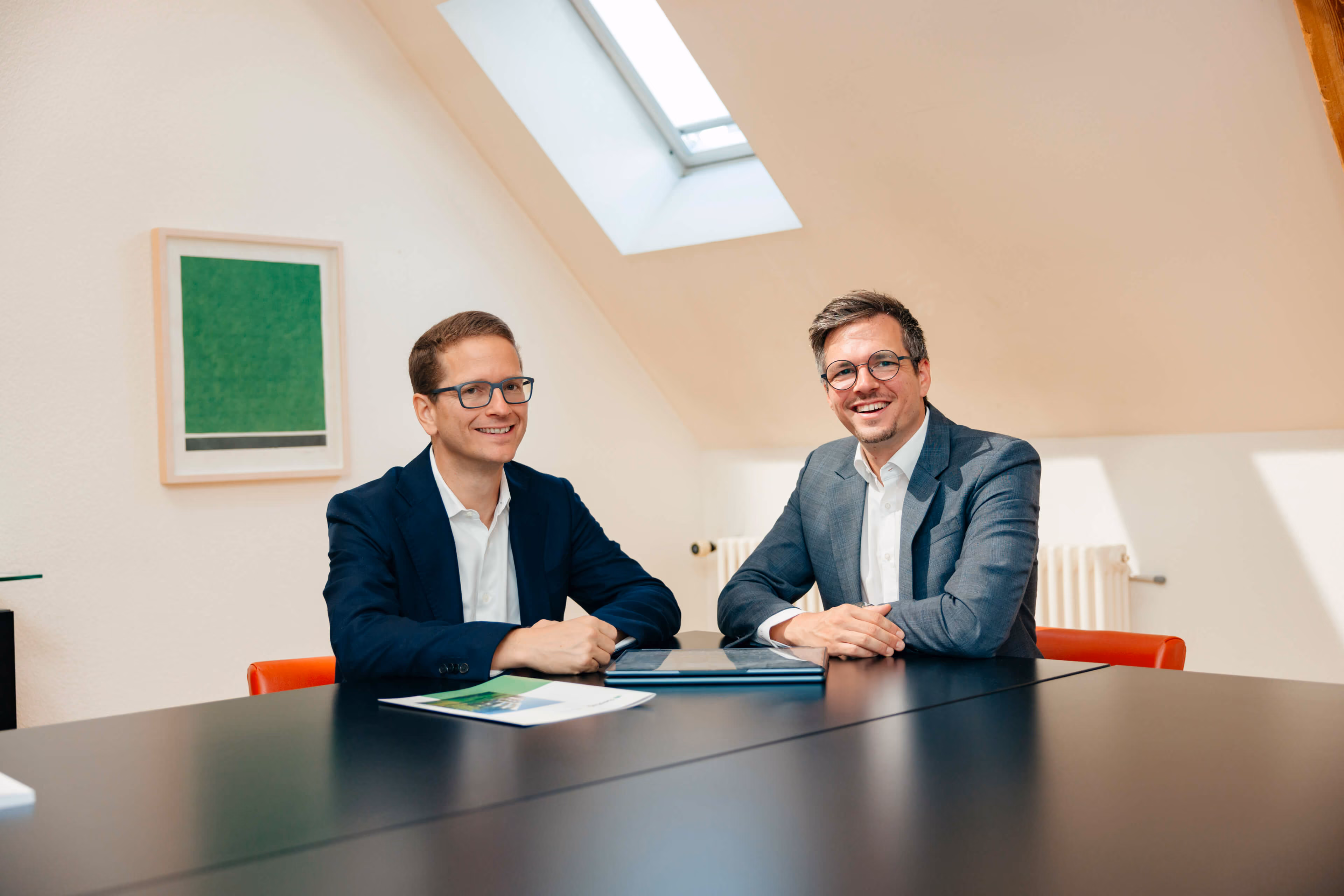Patrice Choffat und Gabriel Diezi in Anzügen sitzen an einem schwarzen Tisch in einem hellen Raum mit Dachfenster und lächeln in die Kamera, auf dem Tisch liegen Unterlagen und ein geschlossenes Laptop.