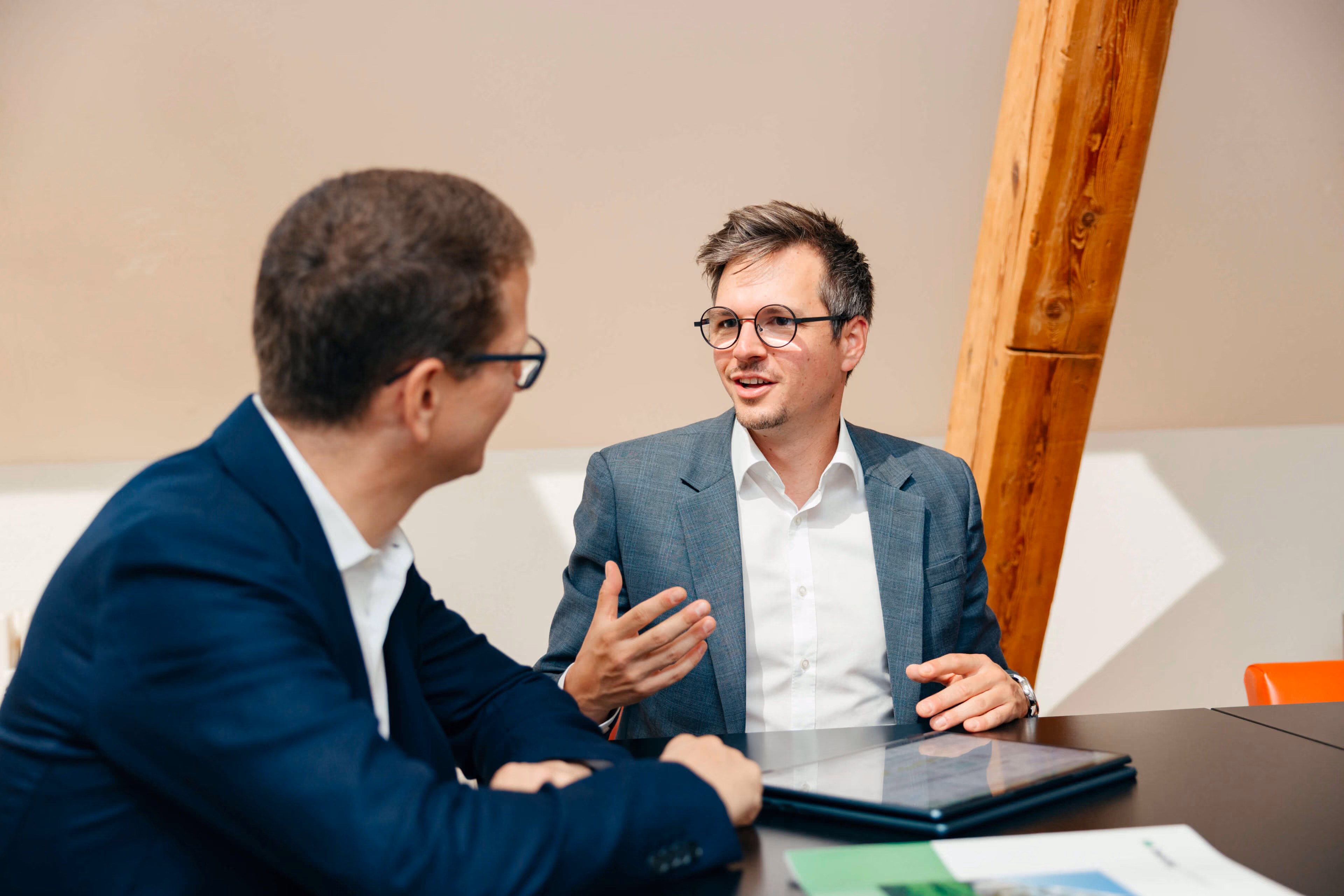 Patrice Choffat und Gabriel Diezi in Anzügen sitzen an einem Tisch und führen ein Gespräch, einer von ihnen gestikuliert, vor ihnen liegt ein Laptop und ein Dokument.
