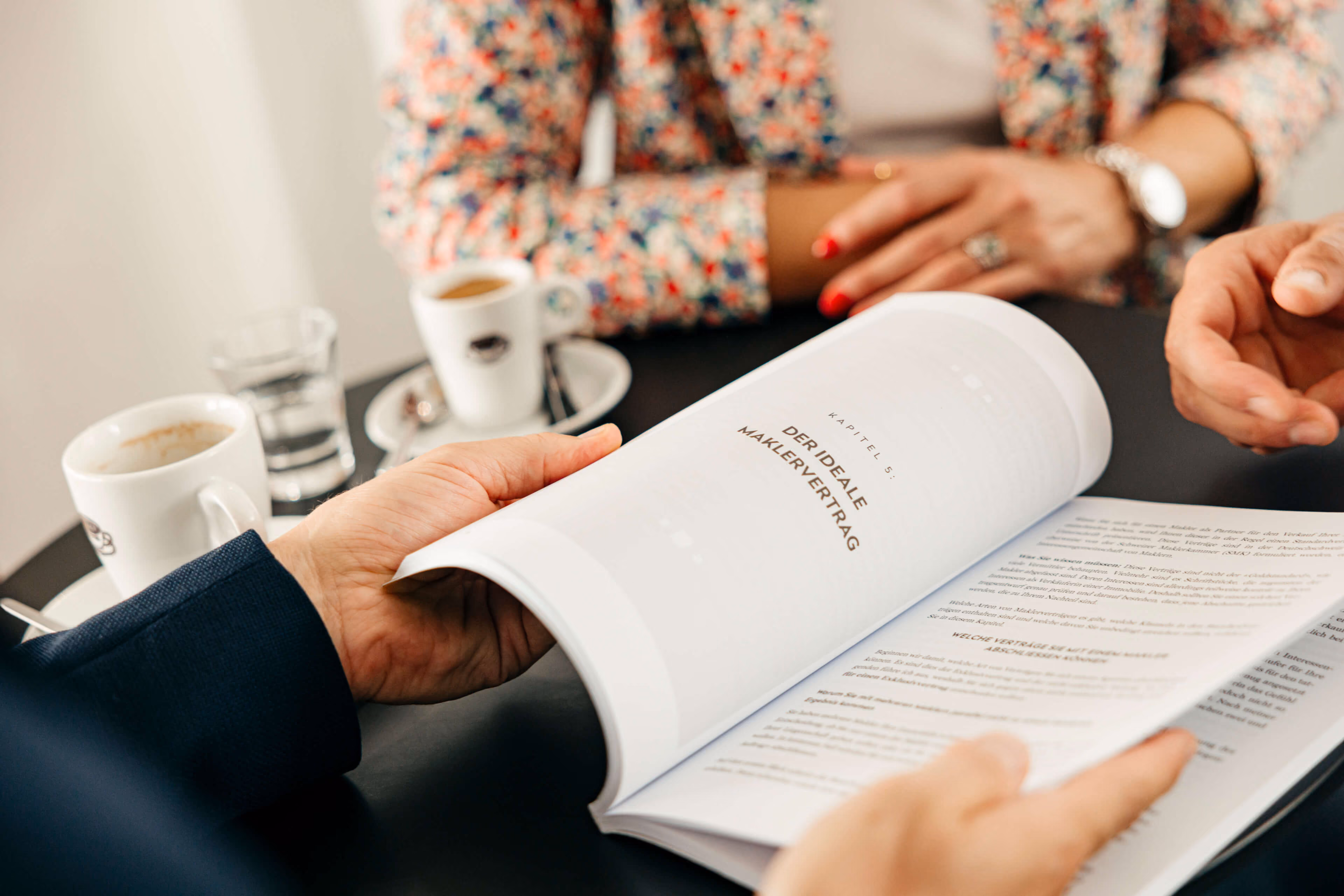 Zwei Personen sitzen an einem Tisch mit Kaffees, eine hält einen Vertrag mit dem Titel "Der ideale Maklervertrag" in den Händen.