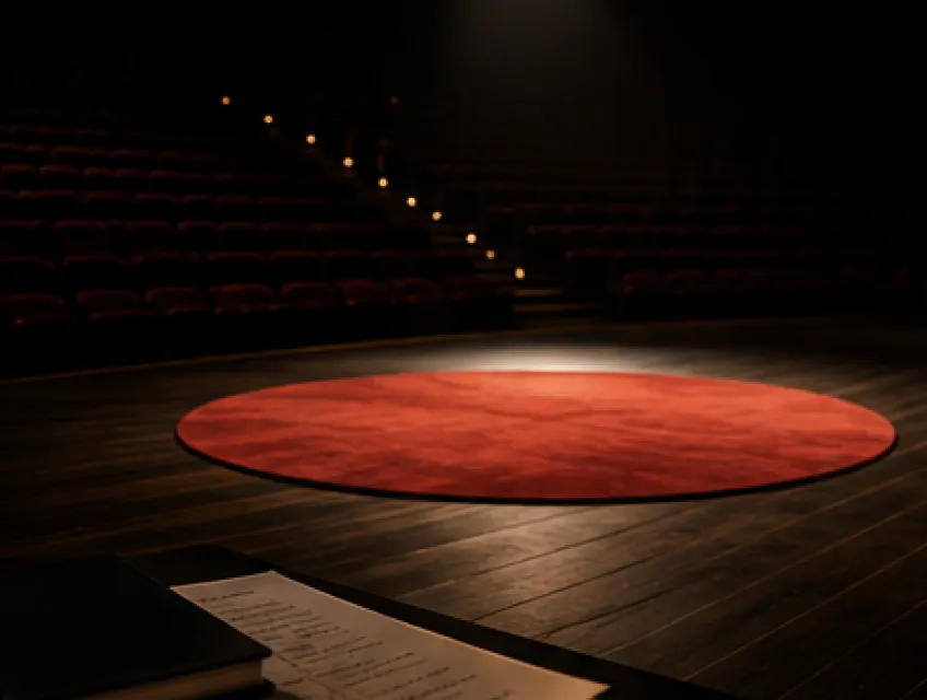 Empty stage with a round red carpet under a spotlight and a dark auditorium with stairs lit by small lights in the background. A notebook and paper are visible in the foreground.
