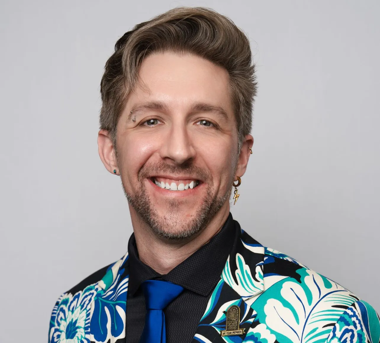 Smiling man with light brown hair styled up, wearing a blue and white floral patterned jacket, black shirt, and blue tie, with earrings and a pin that reads '25 Year Member'.